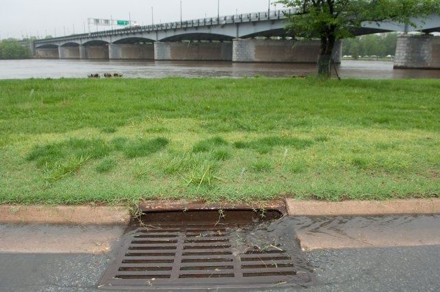 Roadside storm drain beside a grassy riverbank and bridge